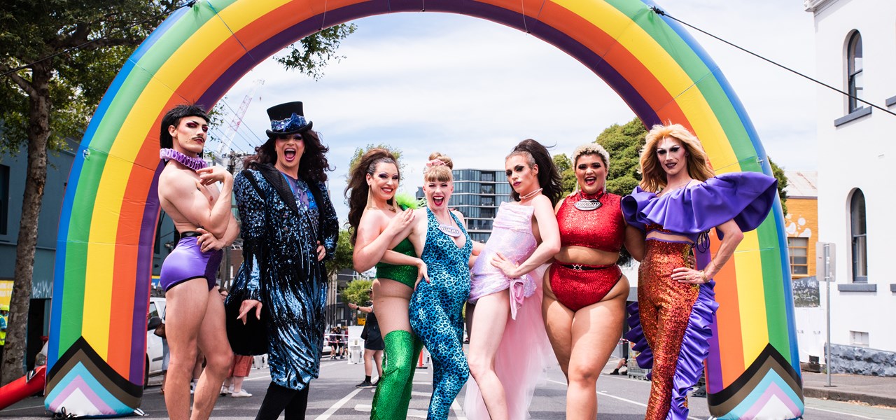 Seven performers stand in-front of a large blow up rainbow for pride