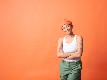 Comedian Aurelia, wearing a white top and green pants, is standing in front of an orange background.
