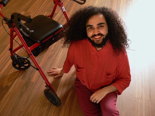 Patrick sitting on the floor, dressed in red tones, with a wheelable walker frame behind them. Patrick has long, curly black hair and a black beard and moustache.