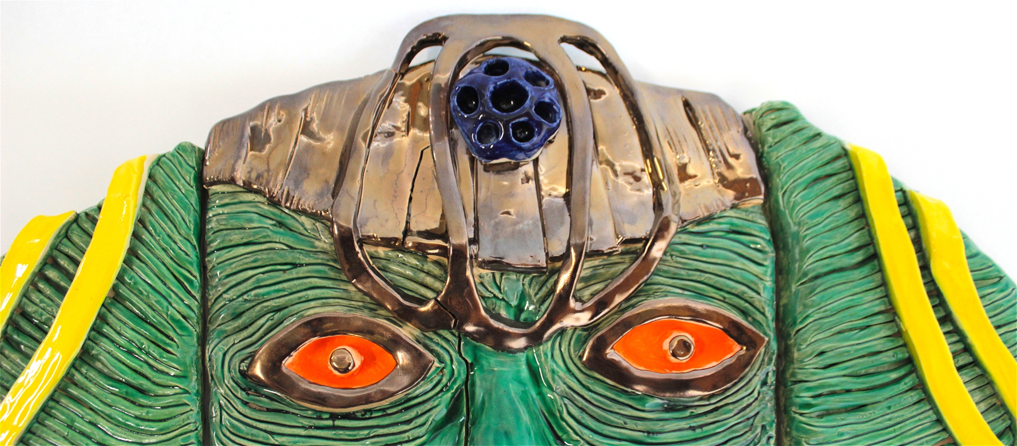 A ceramic head that has green skin, with two orange eyes, a gold head dress and gold beard. There's a blue token on its forehead.