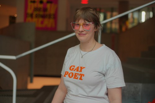 Alex wearing a white t-shirt with orange GAY POET text wearing pink-shaded glasses and a silver necklace