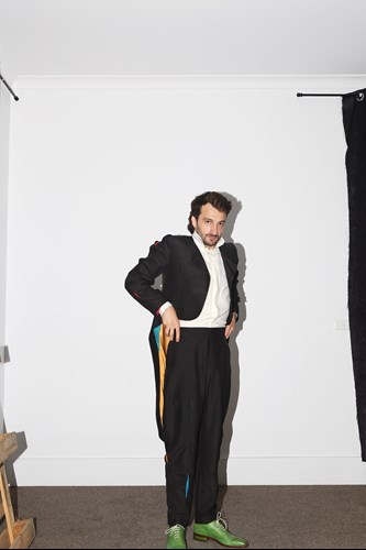 Jacob Abela in a black suit, white shirt and green shoes stares at the camera against a white background