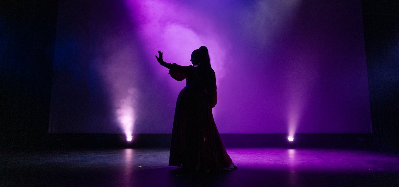 Silhouette of a performer in purple lighting. Performer is wearing a long dress, standing with their right hand out and looking towards it.
