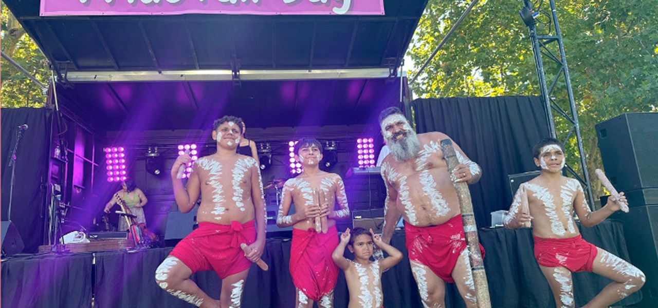 Five indigenous dancers in native attire standing on a lawn in front of a stage with text PRIDE FAIR DAY