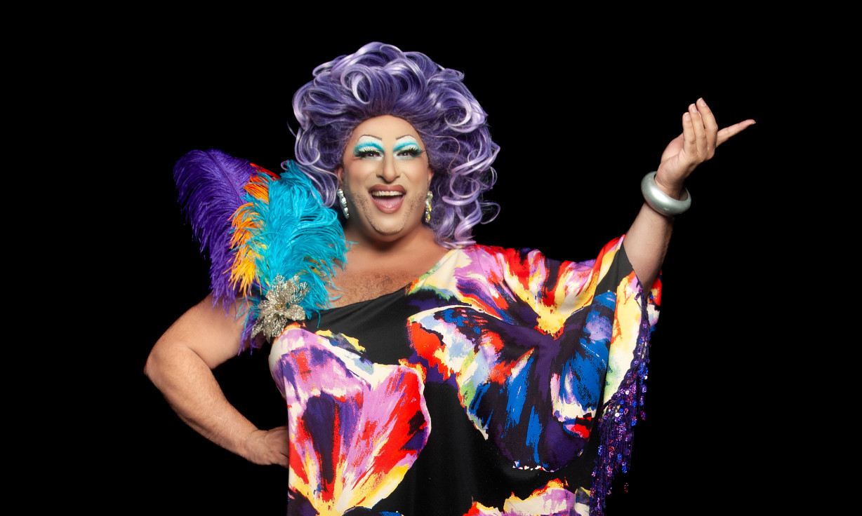 Frock Hudson in a glamorous colourful dress and mauve wig against a black background