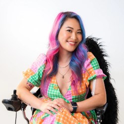 A person dressed in a colourful checked dress with colourful hair sitting in a wheelchair
