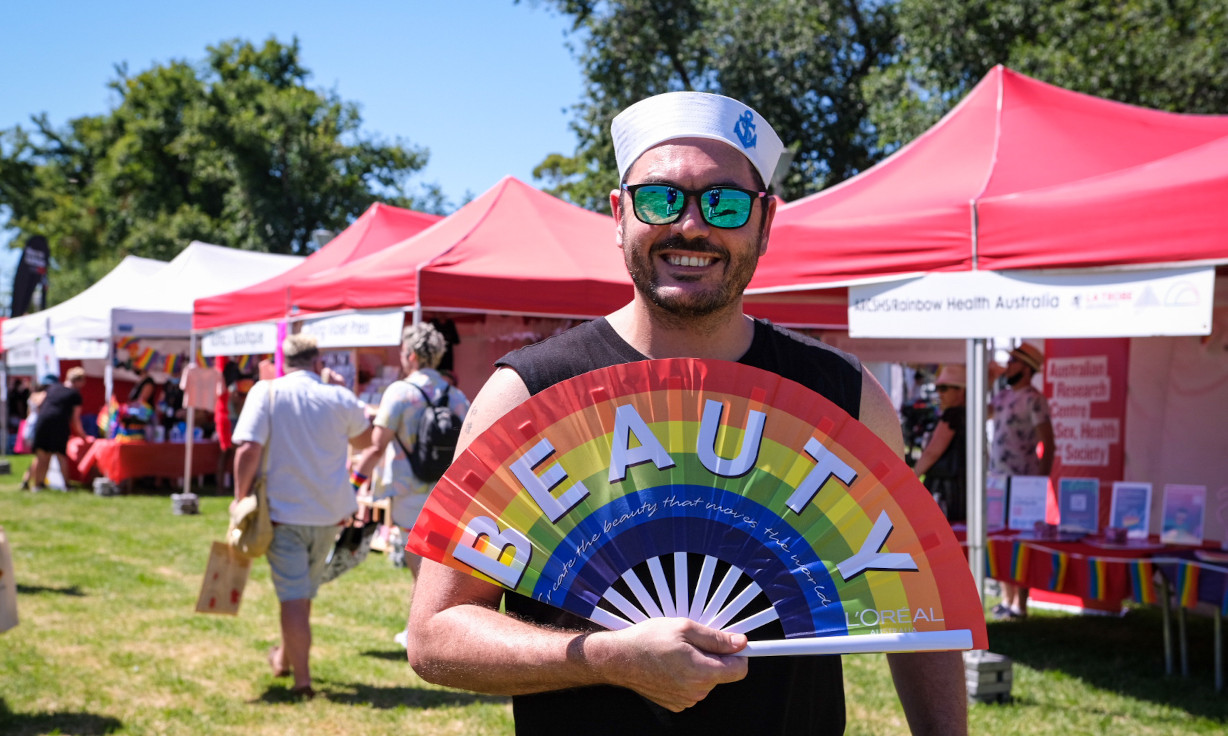 Person wearing sailor hat holding an opened rainbow fan with text "BEAUTY", standing in front of Midsumma Carnival stalls