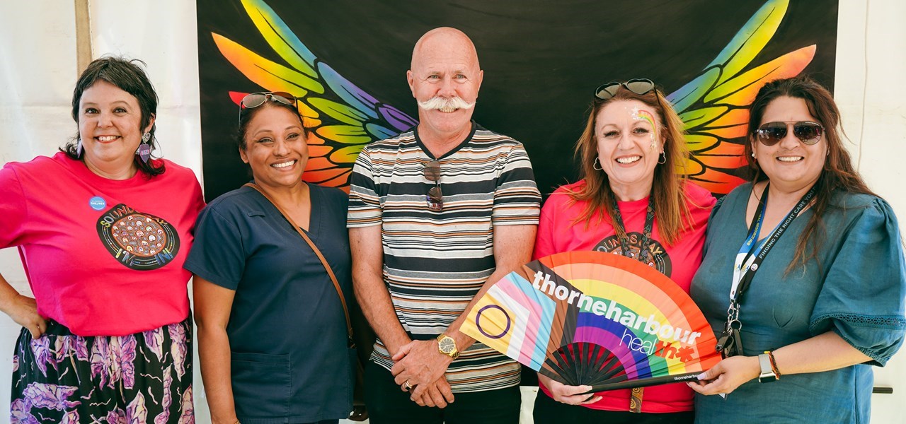 Five people posing for the camera. One is holding a Thorne Harbour Health fan. There is a rainbow bird in the background.