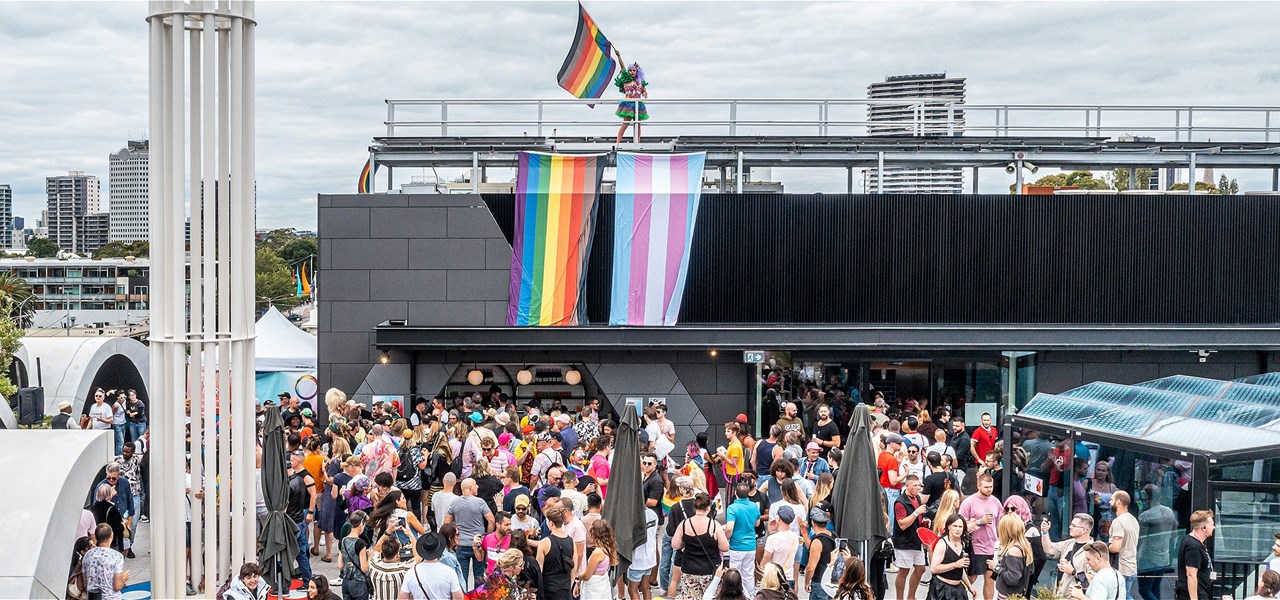 People dancing at a Rooftop party. A progress flag and a trans flag are displayed side by side. A drag queen is waving a progress flag on the roof.