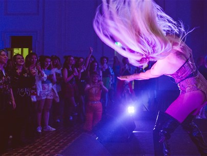 There is a drag performer dancing on a low stage whipping their long blonde hair around, being cheered on by dancing young people in a hall.