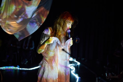 Photo of a person with long hair singing into a microphone in front of a black background but with clothing and background fabric colourfully lit