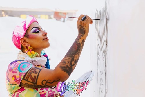 Cynthia is wearing a pink hat and a bright multicoloured shirt, holding a paintbrush in one hand and a palette in the other as they paint on a large canvas.